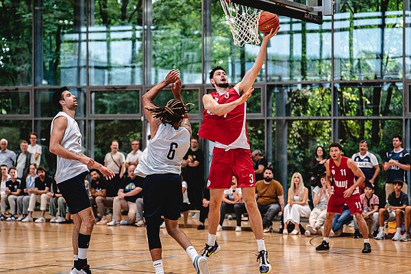 Das Foto zeigt Baskets-Center Leo Saffer beim erfolgreichen Korbleger gegen zwei Heidelberger Verteidiger. Im Hintergrund sind Baskets-Spielmacher Brae Ivey und einige Zuschauer zu sehen.